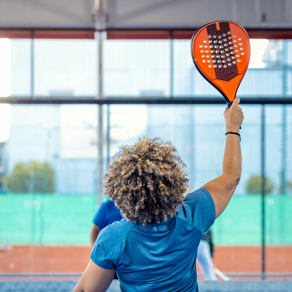 Player serving during an indoor padel match with racket raised toward the ball