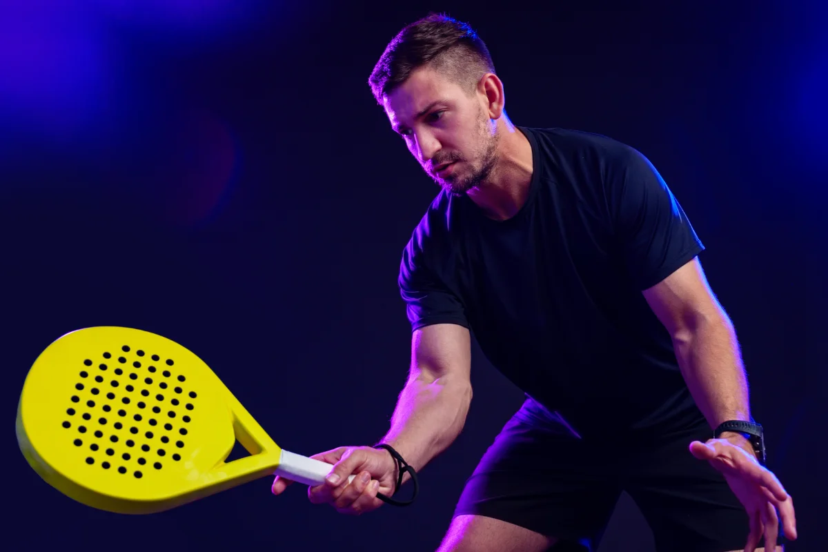 Man playing padel indoors with a yellow racket on a blue court under studio lighting.