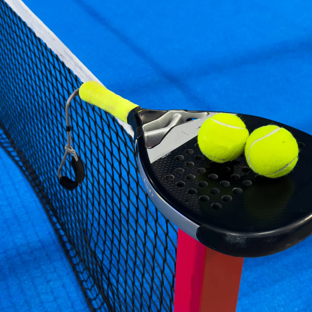 Padel racket balanced on the net with two balls on an indoor blue court