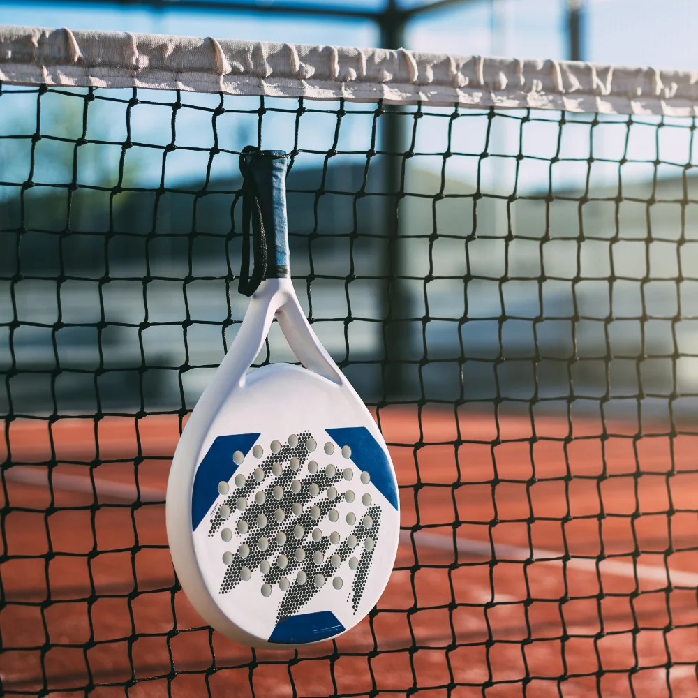 White and blue padel racket hanging on a court net during an outdoor match.